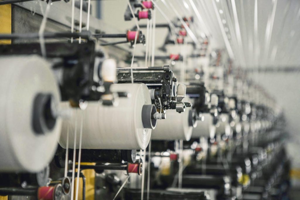 Industrial weaving machines in operation, with bobbins of white yarn lined up in series.