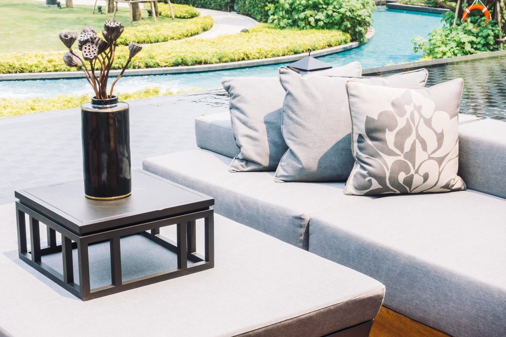 Outdoor living area with a light gray fabric sofa decorated with printed cushions in neutral tones, alongside a black coffee table and a pool in the background, creating a modern and relaxing environment by the water.