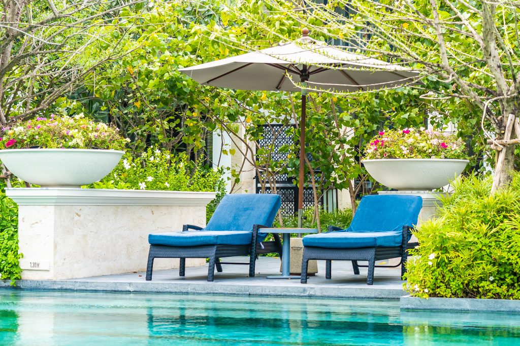 Wicker sun loungers with blue waterproof fabric upholstery, arranged in front of a blue pool, surrounded by green vegetation, in a luxurious and relaxing outdoor environment.