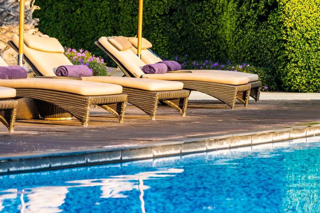 Wicker lounge chairs with beige waterproof fabric cushions, placed beside a blue pool and surrounded by lush greenery, creating a luxurious and relaxing outdoor environment.