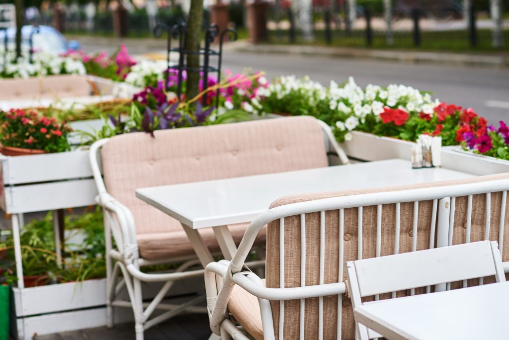 Espaço exterior de um café ou restaurante, com mesas e cadeiras estofadas em tecido bege, rodeadas por floreiras coloridas, criando um ambiente acolhedor e convidativo para refeições ao ar livre.