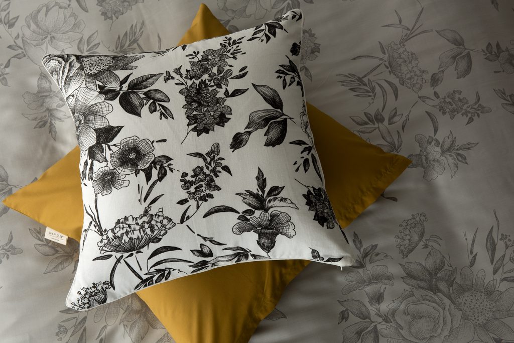 Almofada com estampado floral em preto e branco sobre fundo branco, disposta em cima de outra almofada amarela, num conjunto de roupa de cama com padrão floral.