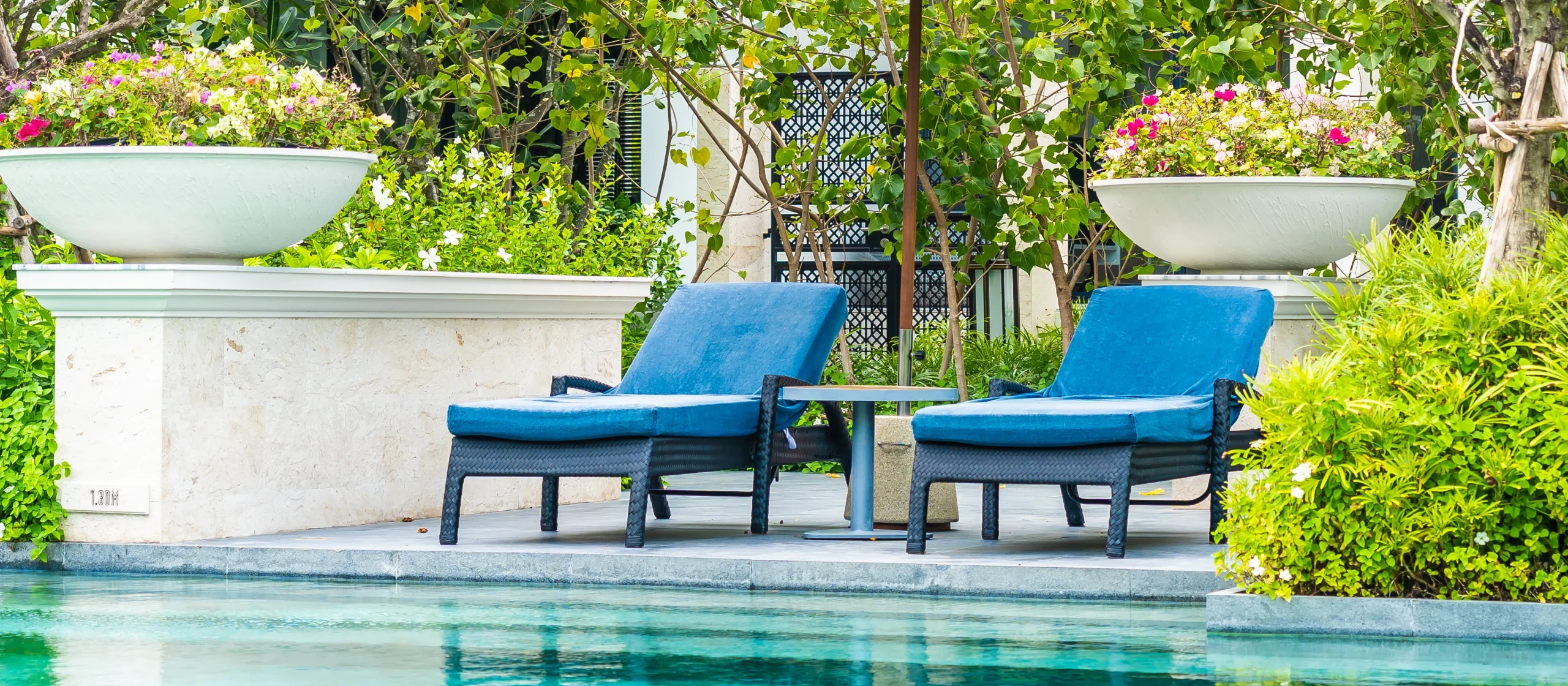 Espreguiçadeiras de vime com estofos em tecido impermeável azul, dispostas em frente a uma piscina azul, rodeadas por vegetação verdejante, num ambiente luxuoso e relaxante ao ar livre.