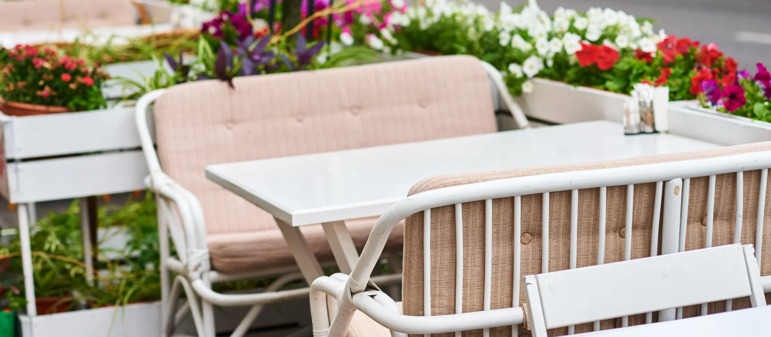 Espaço exterior de um café ou restaurante, com mesas e cadeiras estofadas em tecido bege, rodeadas por floreiras coloridas, criando um ambiente acolhedor e convidativo para refeições ao ar livre.