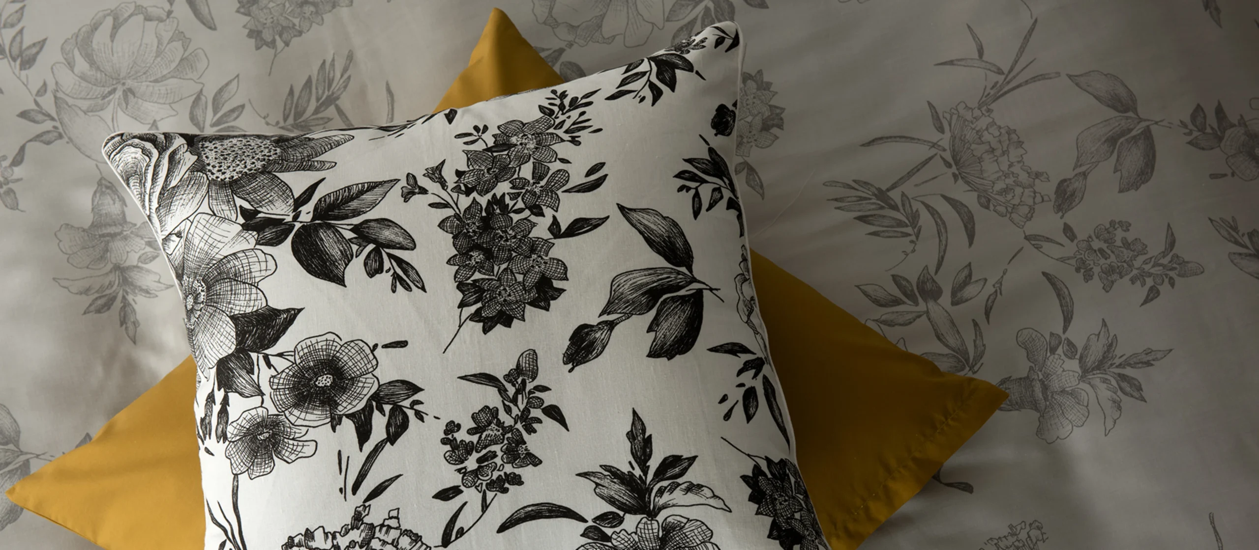 Almofada com estampado floral em preto e branco sobre fundo branco, disposta em cima de outra almofada amarela, num conjunto de roupa de cama com padrão floral.